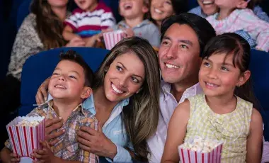 Two smiling parents and their children seated in a theater, enjoying a movie with popcorn in their laps