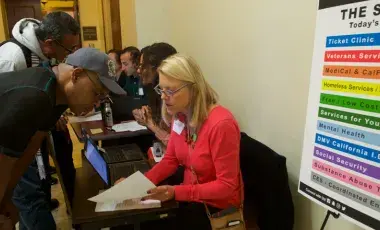 Seated woman in a red sweater at a table helping a standing man with paperwork.