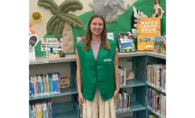 A young woman wearing a green "STAR" vest and a floral skirt standing in a children's library surrounded by books and colorful wall decorations.