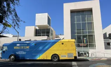 Blue and Yellow Mobile Eye Clinic Bus parked in fron tof a building. The blue sky is in the background.