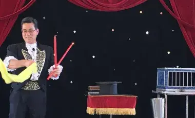 A magician holding wands and a scarf on stage next to a table with a top hat and a cage
