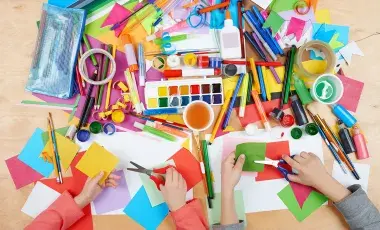 An assortment of craft supplies, paints, markers and hands with scissors cutting paper.