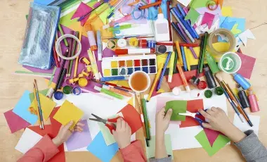 an assortment of paints, markers, paper, glue, scissors and other art supplies shown from above with two sets of hands at the bottom cutting paper.