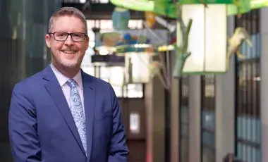 A man in a blue suit and glasses smiles in the Atrium of Los Angeles Central Library.