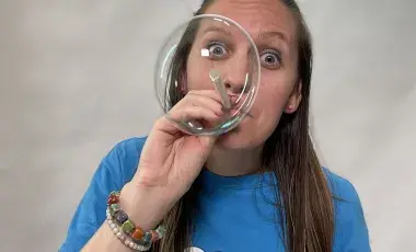 Woman in a Blue shirt blowing a bubble that covers her face. 