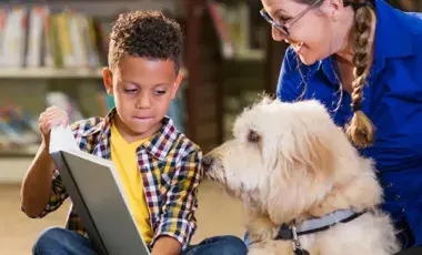 Young boy holding up a book for a shaggy dog to look at.