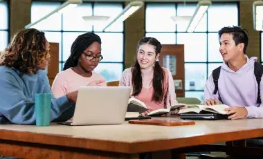 Teens sitting around a table in the library with books and a laptop.