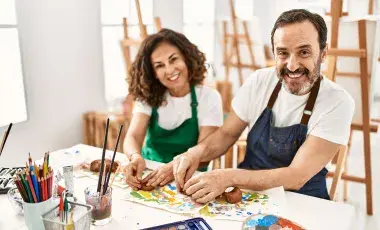 Two adults wearing aprons are smiling and working clay with their hands. The table in front of them has many different types of art supplies.