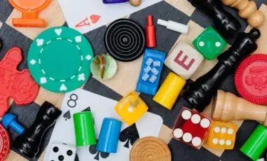 Colorful board-game pieces, cards, dice, chips, pawns, and wooden tokens on a tabletop on a checkered background.