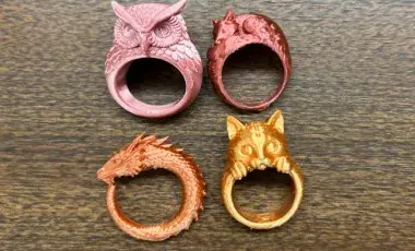 Four 3D printed rings: a pink owl, a burgundy fox, an orange dragon, and a golden cat resting on a counter.
