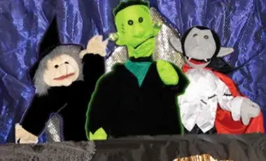 Halloween puppet scene with a smiling witch, green Frankenstein monster, and gray-faced vampire in costumes posing against a purple backdrop.