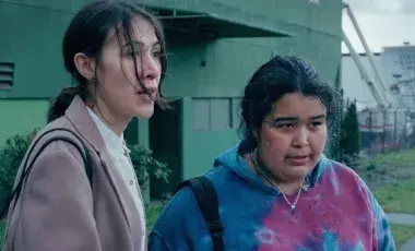 Two indigenous women, one wearing a tie-dye sweatshirt and the other in a more upscale tan coat, stand on a Vancouver street