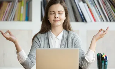 A young woman sitting at a desk with a laptop open. The young woman has her eyes closed and her hands out to her side as if in a meditative state.