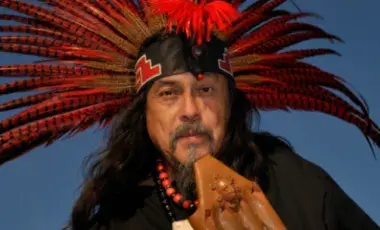 Performer Martin Espino holds an instrument and wears a feathered red headdress.