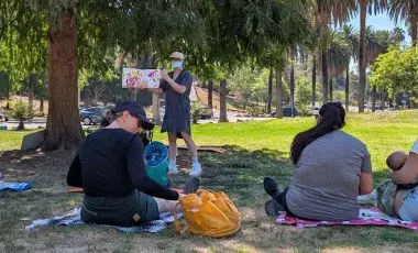 masked librarian reads a picturebook to a group of adult caregivers and young children on a lawn under a shady tree