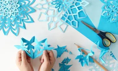 Hands hold a paper cut snowflake with blue snowflakes, scissors and paper on a table.