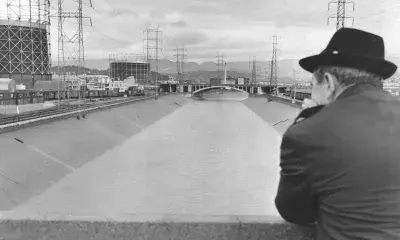 Person in overcoat and wide-brimmed hat facing away from the camera, looking down at the Los Angeles River with industrial tanks in the background.
