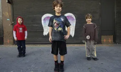 Three children pose in front of rollup storefront door, with the center child in front of wings