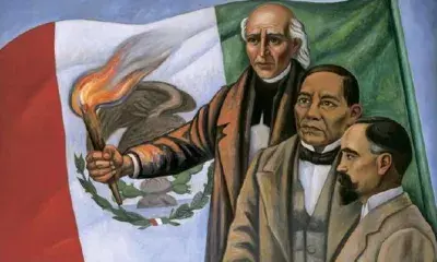 Three people stand in front of a Mexican flag, one holding a torch