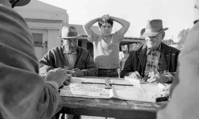 Young boy stands over outdoor table with men looking at a newspaper