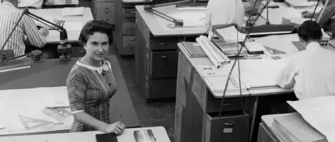 Newspaper employee standing while other employees sit and work at desks.