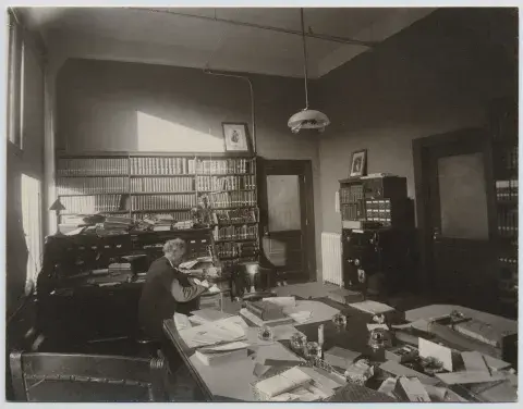 Charles Lummis sitting at his desk with a pen in hand. The windows are behind him to the left and the doors are on the right.
