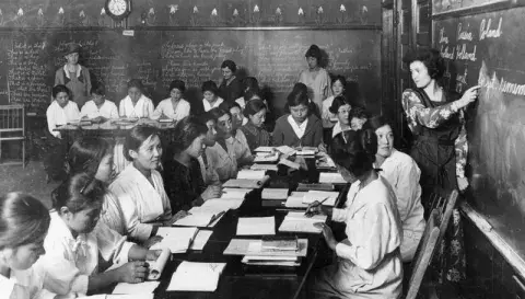 Students around a table watching the instructor point at the chalkboard.