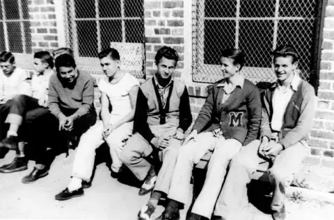 A group of students seated on a bench outside near a building.