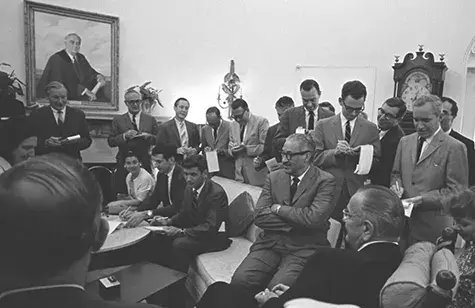 President Lyndon Johnson seated at the end of a couch surrounded by a group of reporters writing notes.