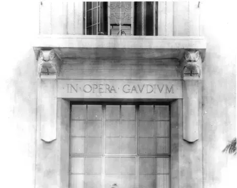 Iron balcony with two sets of stone owls below it and the inscription "In Opera Gaudium."