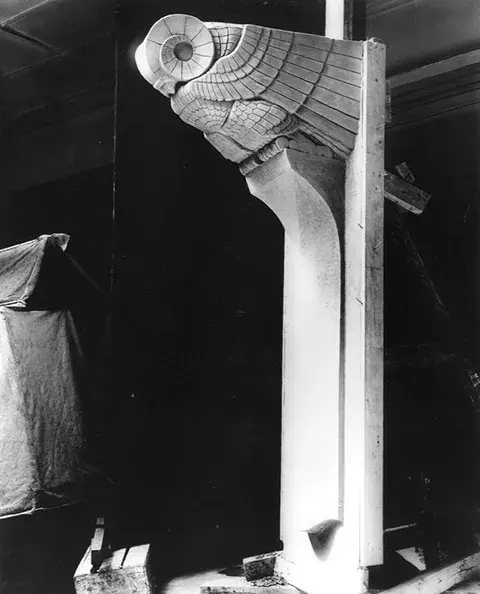 Side view of one stone owl at the top of a column. The body is covered by its wings. The head features spiral patterns.