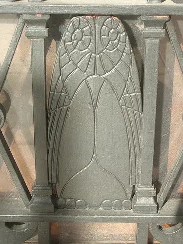 An owl metalwork on a gate. The body is covered by its wings. The head features spiral patterns.