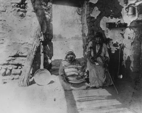 Two elderly women sit in front of an adobe house