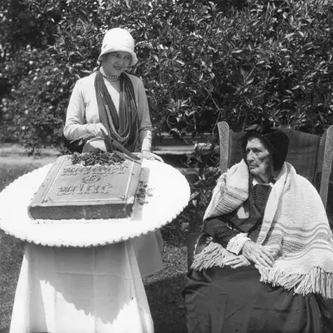 very aged woman is presented a large cake decorated in the shape of a book entitled, "Mother Mine," on Mother