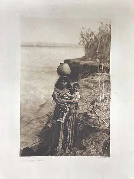 a mohave woman carrying a jug of water on her head while holding a toddler