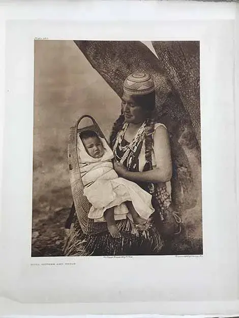 A human woman holding a Hupa toddler. The toddler is wrapped and swaddled.