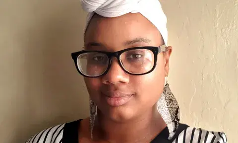 Diamond Forde wearing glasses and a head wrap, leaning against a wall