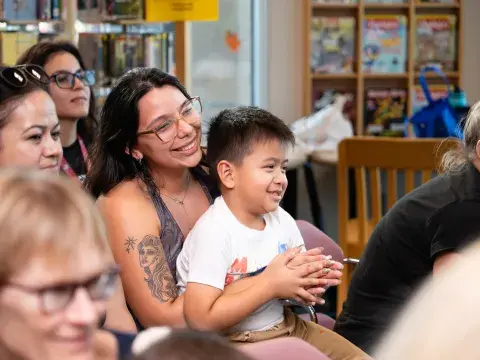 A woman wearing glasses sits in an audience with a young child in her lap. Both are smiling and watching something out of the frame.