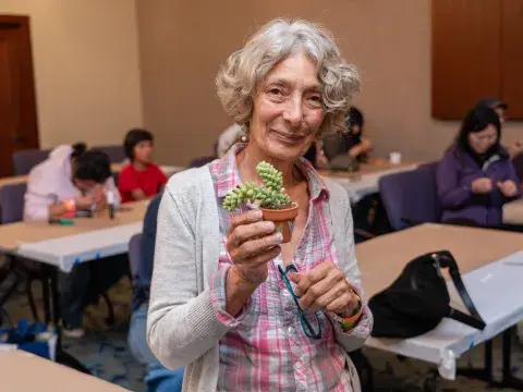 A woman in a cardigan and plaid shirt holds up a succulent in a clay pot in a classroom setting.