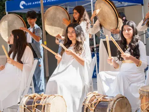 Three teenage girls in white gowns hold large drumsticks above large, ceremonial drums.