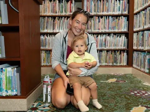 A woman sits on the floor amid library bookshelves with a toddler. 
