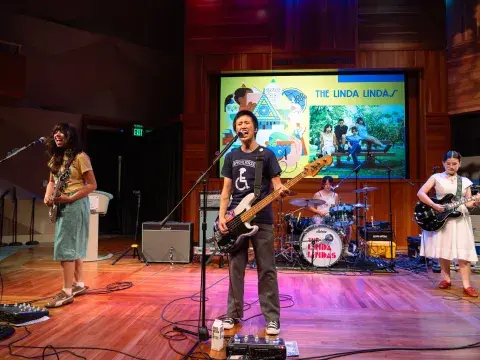 Four rock musicians play on a stage. In the foreground a guitarist plays and sings into a microphone.
