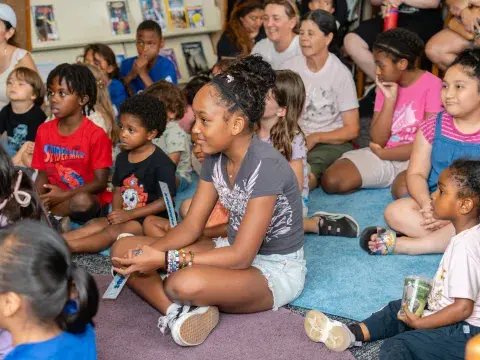 A young girl is seated with other children on a carpeted floor in a library. All are paying attention to something out of the frame and smiling.