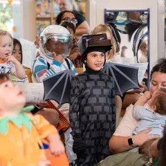 A chile in a bat costume surrounded by children and parents dressed in halloween costumes at a library event.