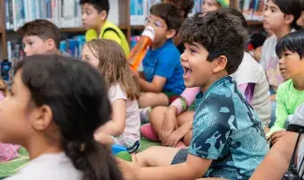 A group of children sit on the floor in a library, watching something with enthusiasm.