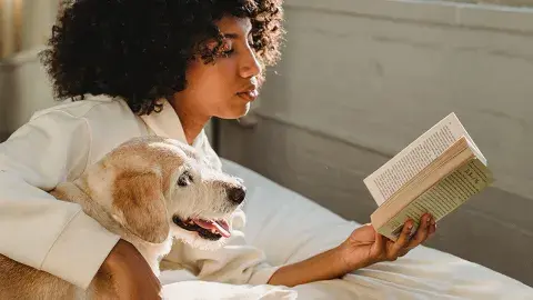A young woman reads a book while her dog lie beside her.