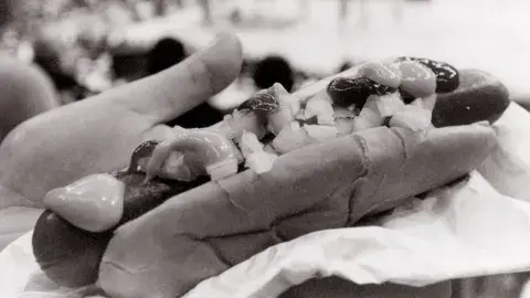 An oversized hot dog fills the bun which is topped with mustard, ketchup and onions.