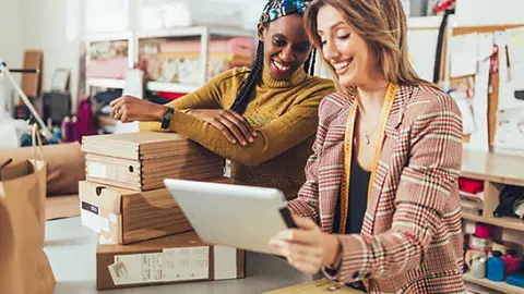 A black and a Caucasian woman are standing next two each other reviewing something in a tablet.  They are in an office with boxes, materials and other tools.