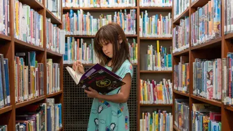 Standing girl, reading a book, surrounded by bookshelves
