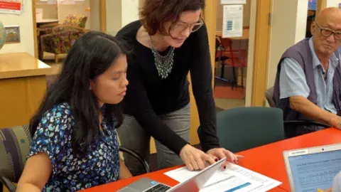 Multiple adults sitting at or standing near a desk, two with laptops, one with a sheet of paper